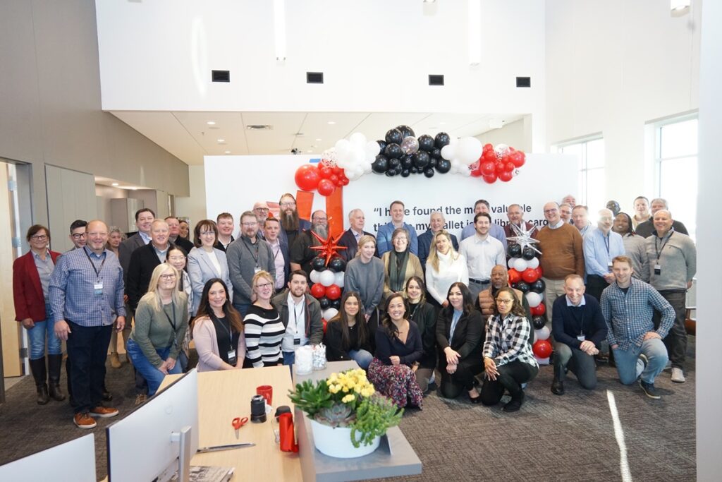 A large group of Bibliotheca employees poses together in front of a red, black, and white balloon arch during the ribbon-cutting ceremony at the new office.