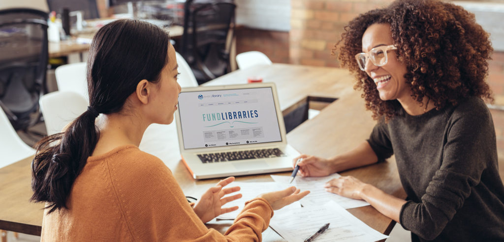 two ladies talking with funding libraries webpage on laptop