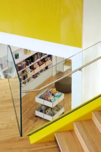 Accès élargi 5 Vue intérieure moderne de la médiathèque B612 avec rayonnages de livres jeunesse et escalier aux finitions bois et jaune.