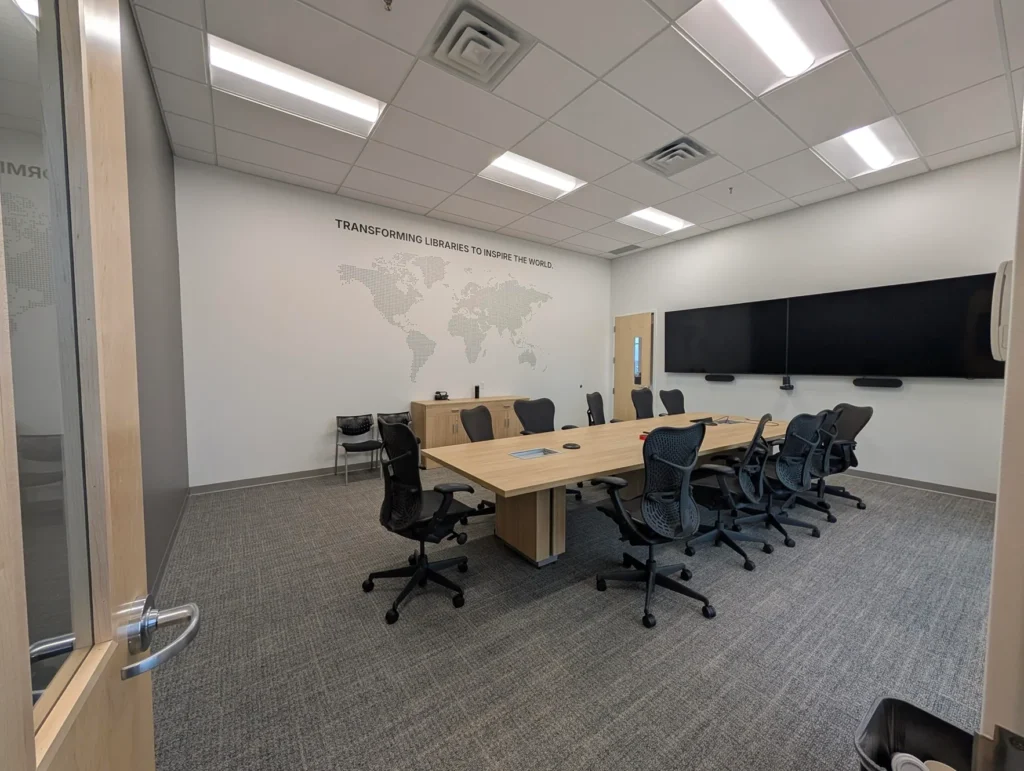 Bibliotheca Opens New Headquarters in Lake Elmo, MN 2 A modern meeting room with a large wooden conference table, ergonomic chairs, and dual wall-mounted screens, featuring a world map decal on the wall.