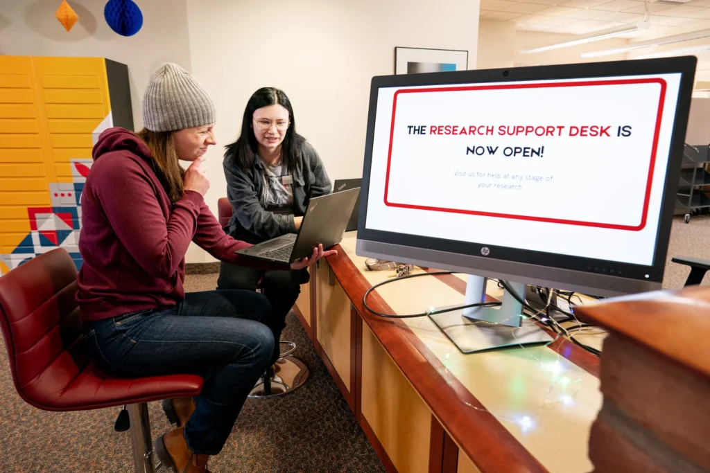 Two librarians collaborate at computers near a sign announcing the new Research Support Desk at Augsburg University’s Lindell Library.